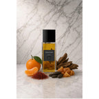 Frasco retangular do perfume Codice d'Oro In The Box, com líquido âmbar, tampa preta e rótulo escuro, sobre superfície clara com fundo de mármore. Ao redor, tangerina, madeira, açafrão e resina âmbar.