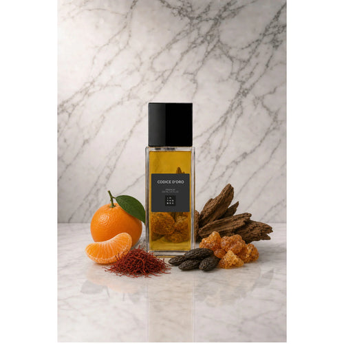 Frasco retangular do perfume Codice d'Oro In The Box, com líquido âmbar, tampa preta e rótulo escuro, sobre superfície clara com fundo de mármore. Ao redor, tangerina, madeira, açafrão e resina âmbar.