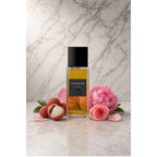 Frasco retangular do perfume Fleur Delicate In The Box, com líquido âmbar, tampa preta e rótulo escuro, sobre superfície clara com fundo de mármore. Ao redor, lichia, rosas e pétalas cor-de-rosa.